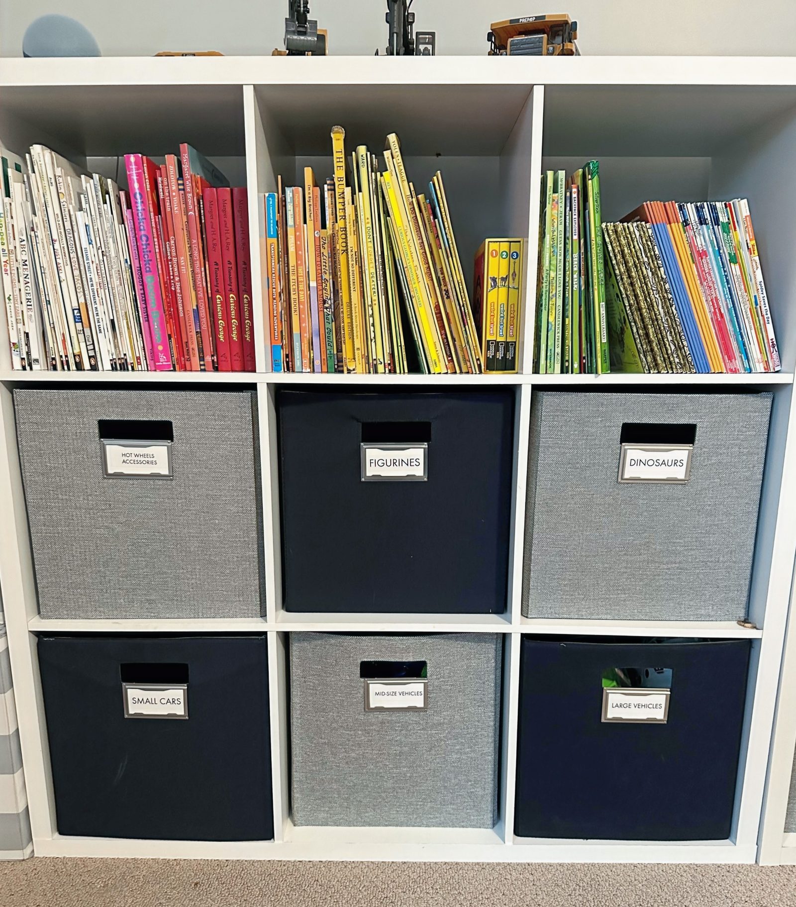 Playroom cube shelves with labeled fabric bins for cars, figurines and dinosaurs, books sorted by color above